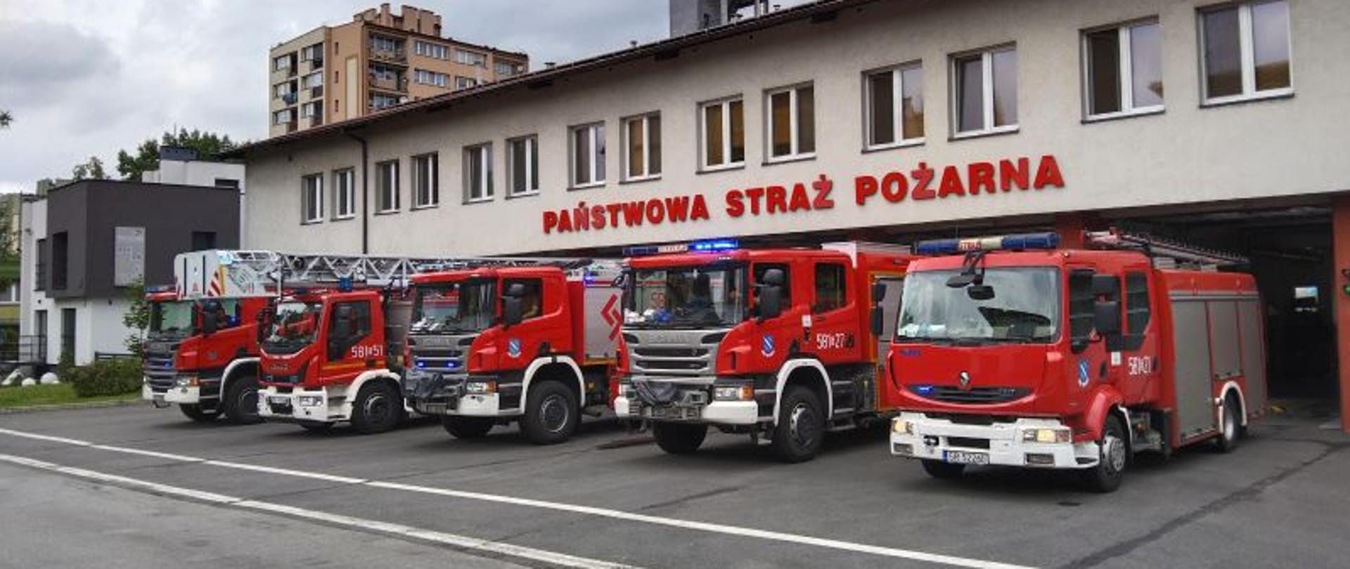 Samochody ratowniczo-gaśnicze przed garażami z włączonymi sygnałami świetlno - dźwiękowymi. 