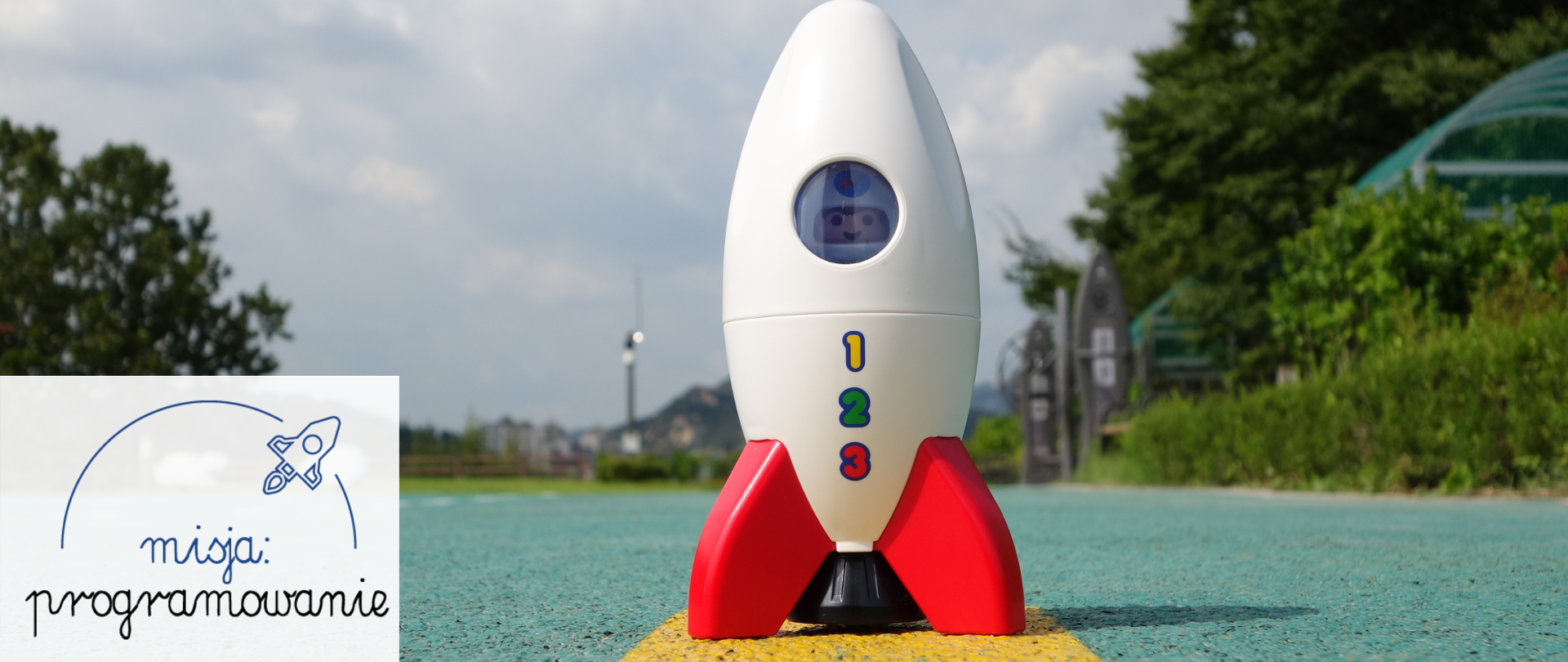 Grafika przedstawia biało-czerwoną rakietę kosmiczną - zabawkę na pasie starttowym z ludzikiem LEGO w środku. W lewym dolnym rogu na zabielonym tle widać napis: misja programowanie.