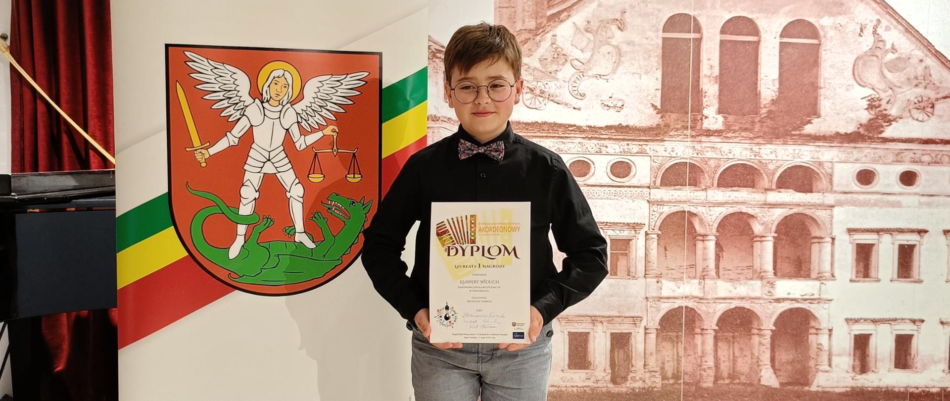 Zdjęcie przedstawia ucznia trzymającego dyplom laureata. Po lewej stronie plakat prezydenta Białej Podlaskiej przedstawiający herb miasta. W tle widoczna ilustracja ukazująca kamienicę.