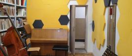 Na zdjęciu przedstawiającym bibliotekę PSM znajdują się regały z książkami, pianino, dwa kontrabasy, pulpit i krzesła, na ścianach są gąbki w kolorach żółtych i czarnych