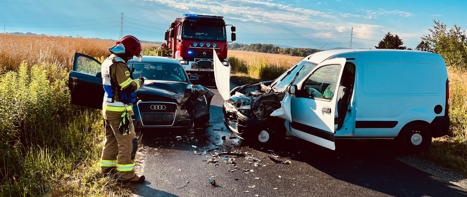 dwa pojazdy osobowe po zderzeniu drogowym, po lewej umundurowany strażak, w tle czerwony pojazd strażacki, wokół pole rolne