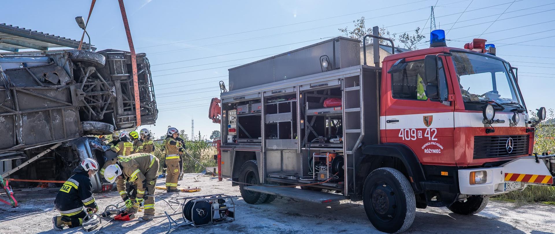 Strażacy prowadzą działania ratownictwa technicznego przy przewróconym na bok ciężkim pojeździe na terenie przemysłowym. Kilku ratowników wykorzystuje sprzęt hydrauliczny i stabilizacyjny w celu wykonania dostępu do osób poszkodowanych znajdujących się pod konstrukcją pojazdu. Nad wrakiem zamocowane są pasy do podnoszenia współpracujące z urządzeniem dźwigowym. W pobliżu znajduje się specjalistyczny samochód ratownictwa technicznego z otwartymi skrytkami sprzętowymi. Na ziemi widoczne są przewody hydrauliczne, narzędzia ratownicze oraz elementy wyposażenia wykorzystywane podczas działań. Zdjęcie przedstawia zaawansowane działania ratownicze prowadzone w warunkach zbliżonych do zdarzenia drogowego z udziałem pojazdu ciężkiego.