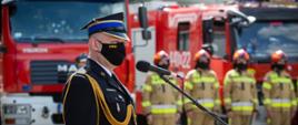 Na zdjęciu widoczny Z-ca Komendanta Wojewódzkiego przemawiający podczas obchodów powiatowych dnia strażaka w Gorlicach. W oddali zmiana służbowa w ubraniach pisakowych specjalnych i czerwonych hełmach w szeregu za nimi czerwone samochody pożarnicze.