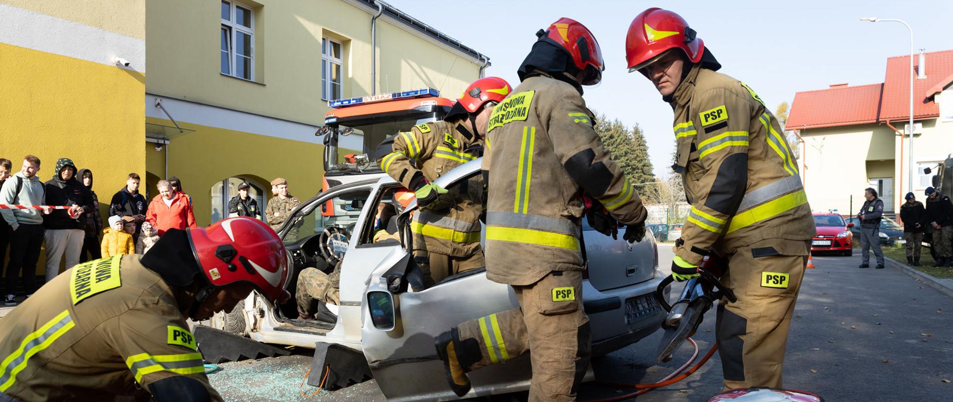 Zdjęcie zrobione na zewnątrz. Na pierwszym planie widać samochód i czterech strażaków. Trzech z nich wycina drzwi z samochodu, a czwarty wyciąga deskę i torbę medyczną. Na drugim planie w tle widać budynek i samochód strażacki.
