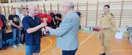 Kolorowa fotografia wykonana w dzień wewnątrz budynku. Przedstawia strażaków na sali gimnastycznej w strojach sportowych i dresach podczas zakończenia zawodów w tenisie stołowym. Na środku Starosta Brzozowski wręczający puchar oraz Komendant Powiatowy PSP.
