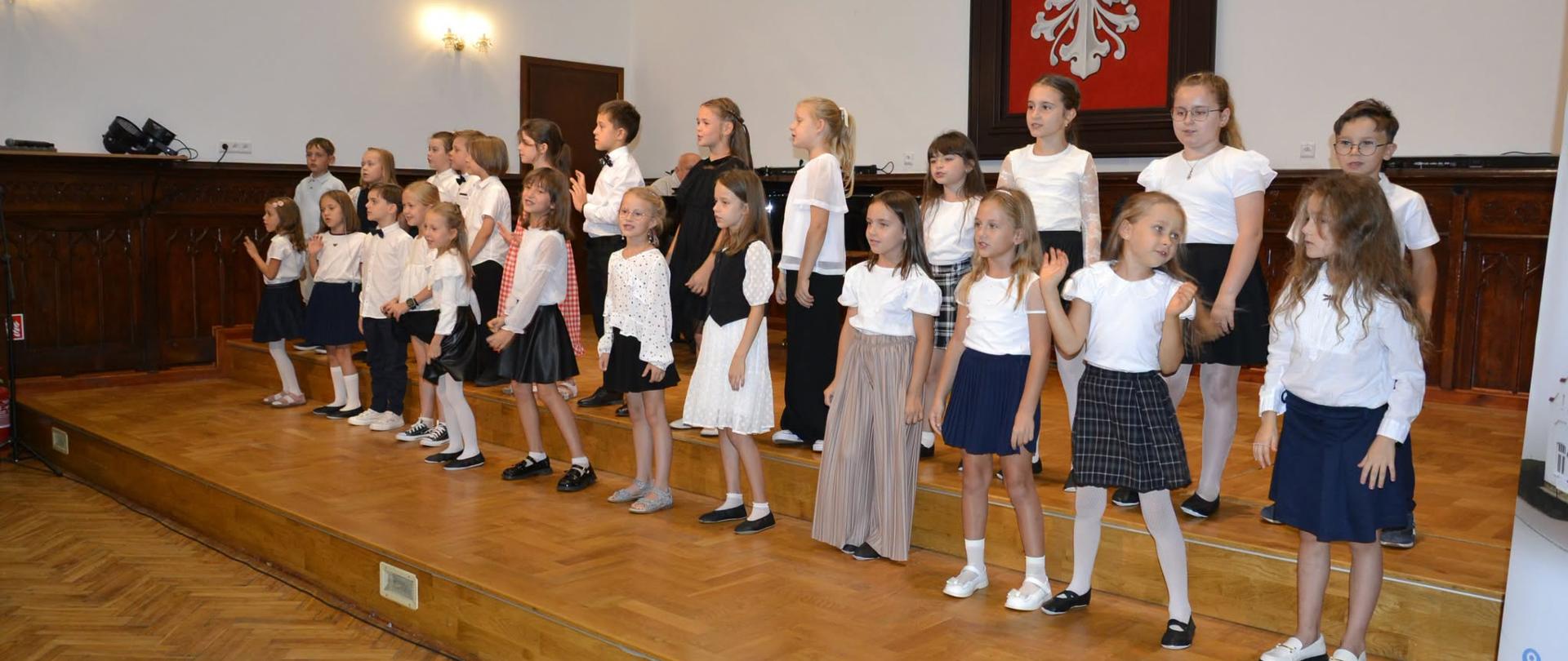 Uczniowie klas pierwszych w białych koszulach i granatowych spódniczkach stoją na scenie Sali Królewskiej podczas uroczystości pasowania na ucznia PSM I stopnia, w tle herb Polski.