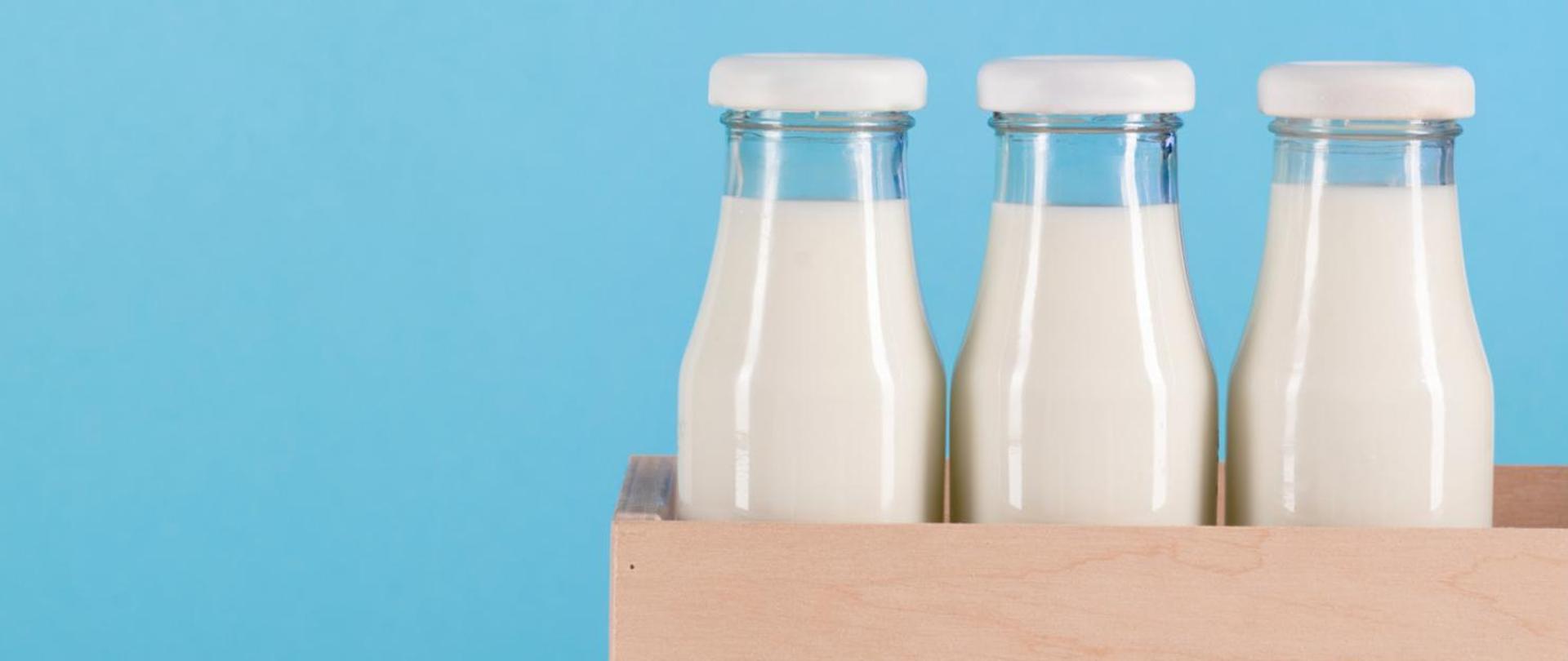 Na zdjęciu są trzy butelki mleka w drewnianej skrzynce. Tło jest niebieskie.