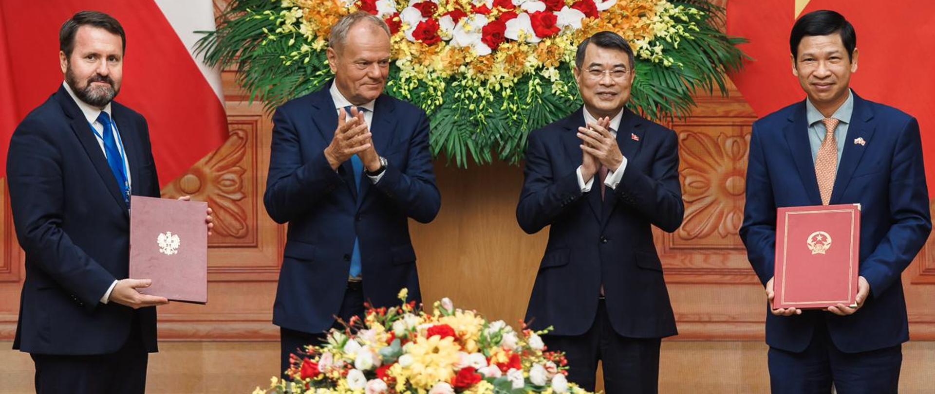 Premier Donald Tusk i Podsekretarz Stanu Robert Kupiecki w Hanoi po podpisaniu umowy o partnerstwie strategicznym. Na zdjęciu znajdują się również premier Wietnamu Lê Minh Hưng wraz z przedstawicielem MSZ Wietnamu. Mężczyźni stoją na tle kwiatów i flag Polski i Wietnamu 