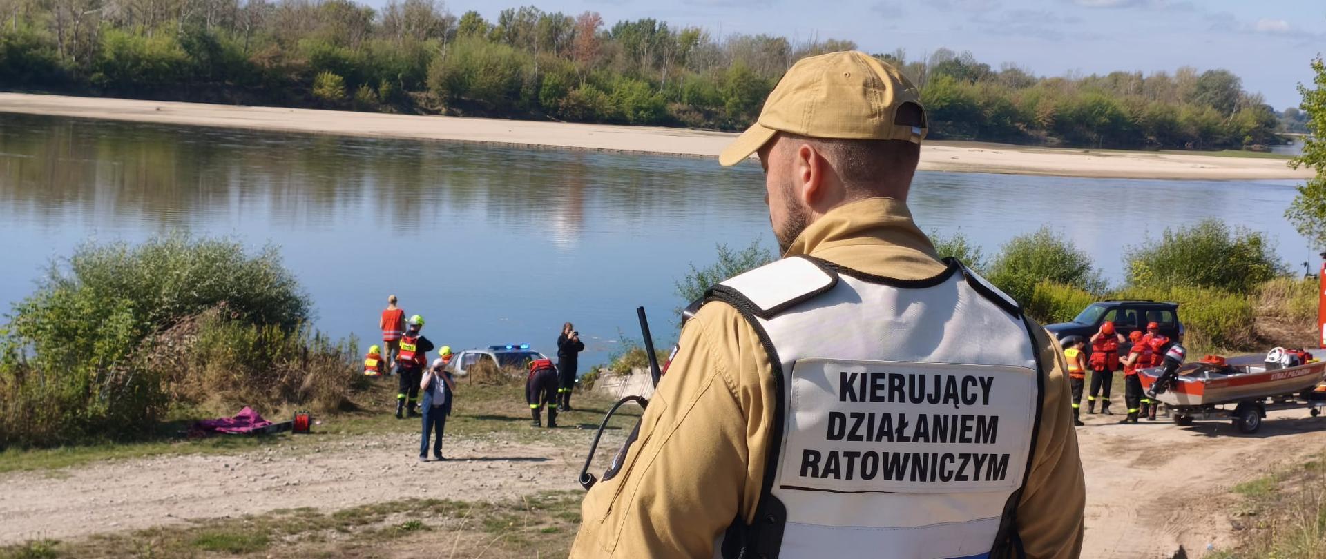 zwarte zdjęcie jest ujęciem zza pleców strażaka ubranego w kamizelkę z napisem "KIERUJĄCY DZIAŁANIEM RATOWNICZYM". W tle widoczna jest rzeka oraz inni strażacy z łodzią.