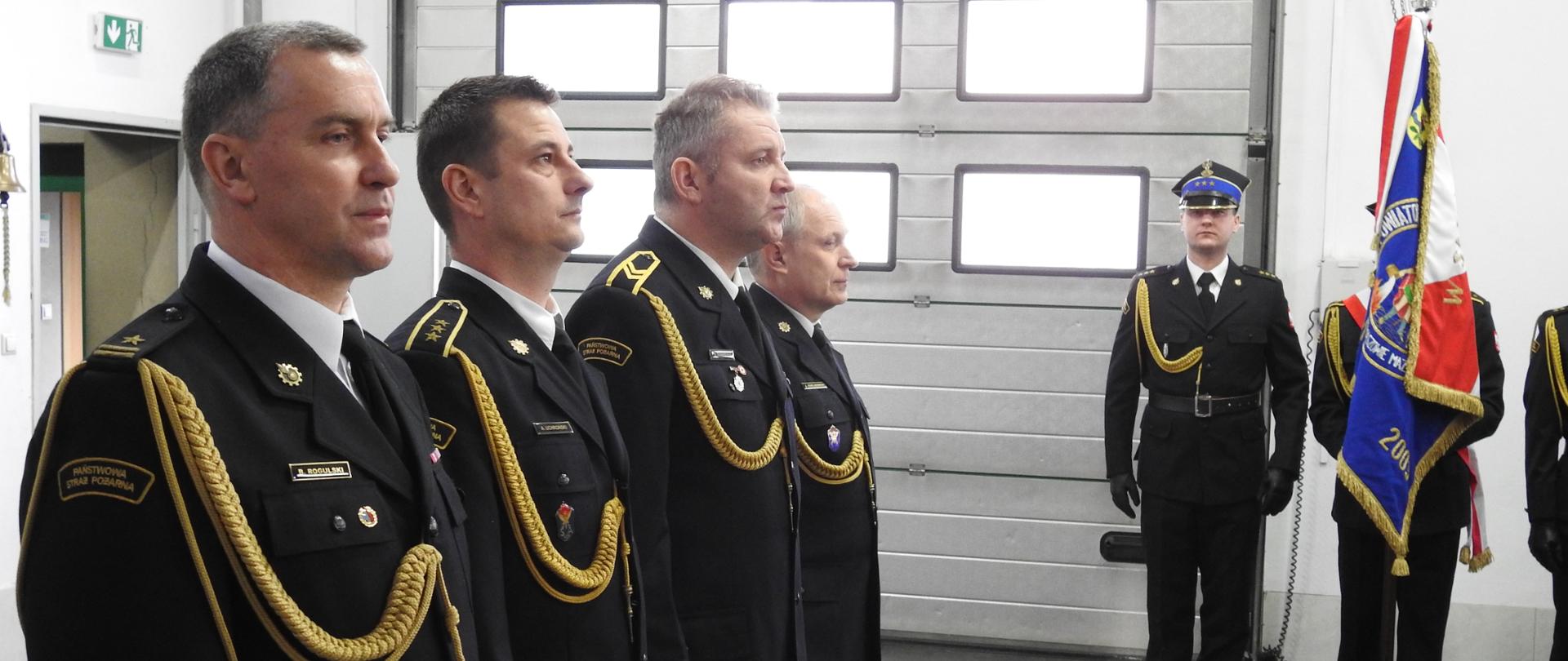 Czterech strażaków w mundurach wyjściowych stoi ustawionych w szeregu. Po prawej stronie na tle białej ściany i bramy garażowej stoi trzech strażak.ów. Strażak stojący w środku trzyma sztandar. 