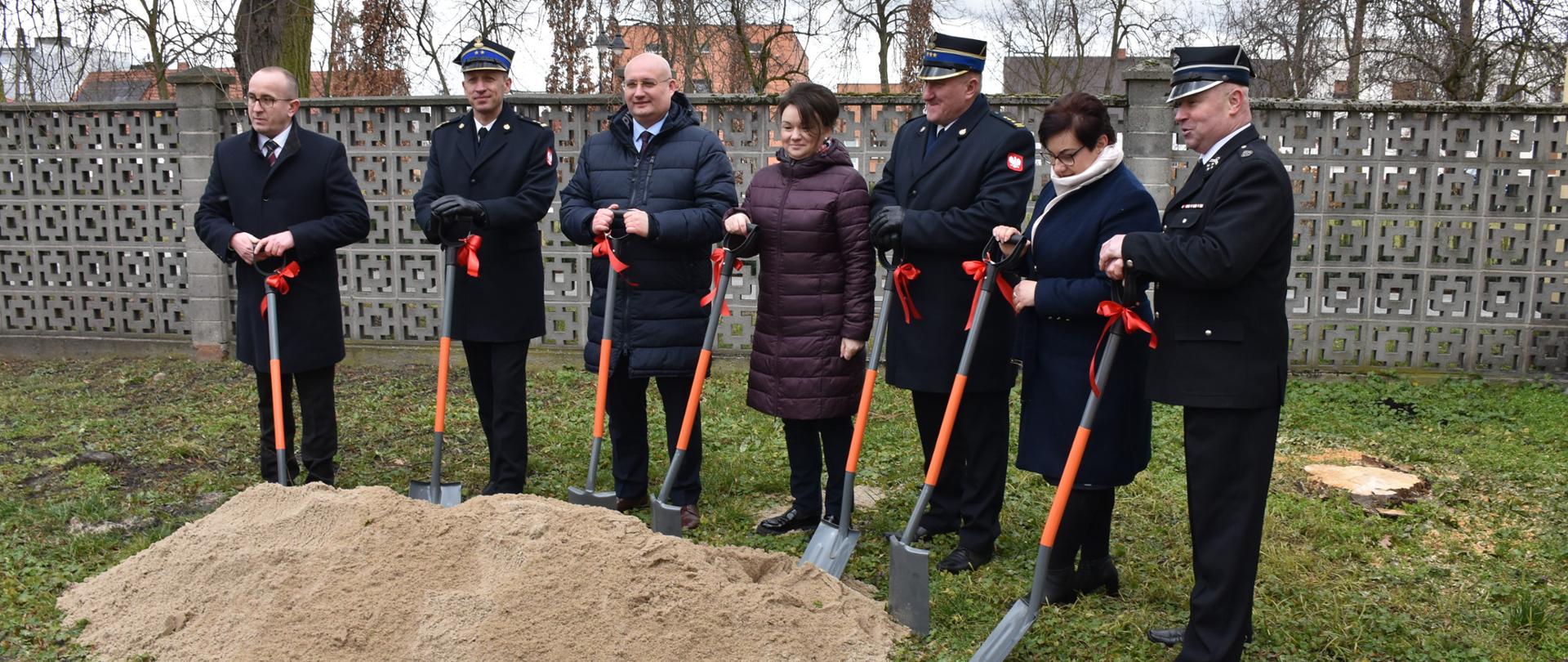 kilka osób w tym strażacy stoją przed dużą ilością ziemi trzymając w ręku łopaty z czerwonymi wstążkami, za nimi szary płot i drzewa