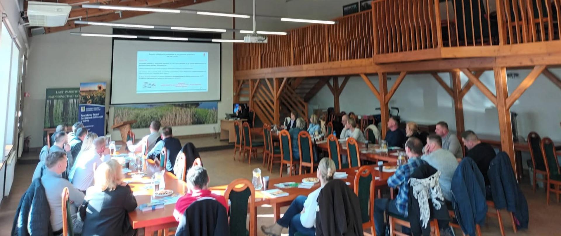 Grupa kilkunastu osób siedzi przy długich stołach w jasnej sali konferencyjnej i uczestniczy w spotkaniu. Na przedniej ścianie wyświetlana jest prezentacja na dużym ekranie. Wnętrze ma wysoki sufit z drewnianymi belkami oraz antresolę z drewnianą balustradą i schodami po prawej stronie. Z lewej strony przez duże okna wpada intensywne światło słoneczne, oświetlając część uczestników.