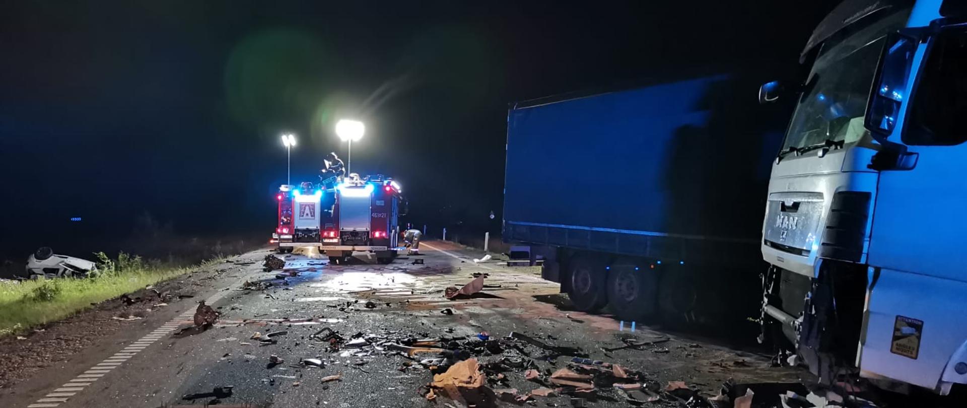 Samochód ciężarowy biorący udział w wypadku stoi w nocy na drodze, w tle dwa samochody pożarnicze.