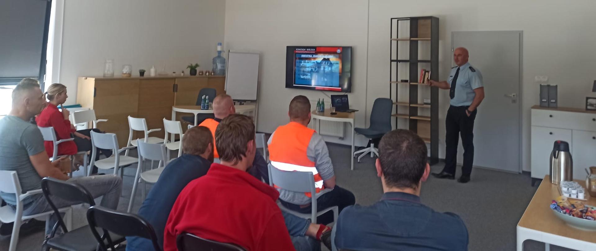 Zdjęcie przedstawia strażaka, który przeprowadza pogadankę na temat bezpieczeństwa w zakładzie pracy. Strażak przy pomocy prezentacji multimedialnej przedstawia zagrożenia występujące w miejscu pracy. Słuchaczami są pracownicy zakładu. 