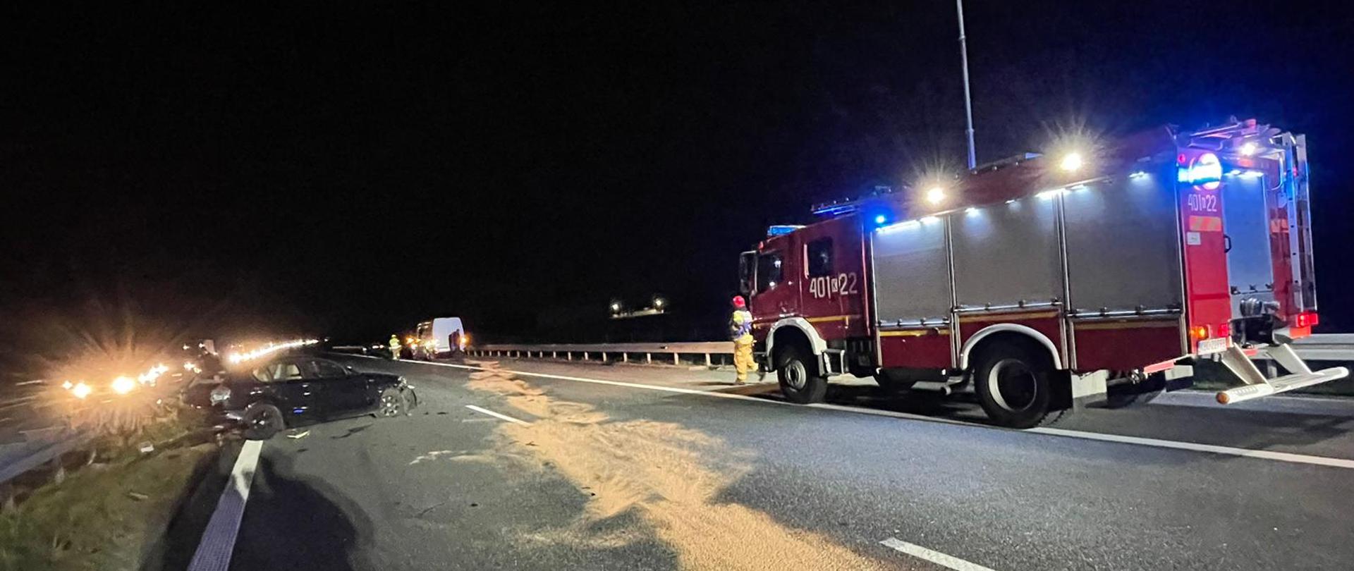 Zdjęcie przedstawia wypadek trzech samochodów na autostradzie A4 w miejscowości Rzezawa w kierunku Krakowa. Na zdjęciu widać na lewym pasie przy barierkach stoi uszkodzony czarny pojazd marki BMW. Po prawej stronie widać czerwony pojazd pożarniczy z włączonymi światłami błyskowymi oraz światłami na zabudowie i dodatkowo ma wysunięty masz oświetleniowy. Przed pojazdem stoi strażak ubrany w piaskowe ubranie specjalne na głowie ma hełm w kolorze czerwonym oraz białą kamizelkę KDR. W oddali po prawej stronie widać uszkodzony biały samochód typu bus. Na drodze widać rozsypany sorbent 