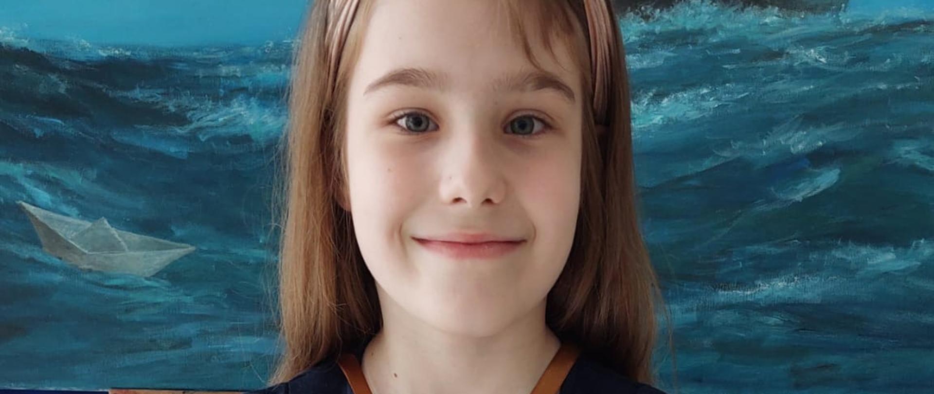 Dziewczynka stoi trzymając dyplom Brązowego Medalu w rękach, na szyi ma zawieszony brązowy medal w kształcie klucza wiolinowego.