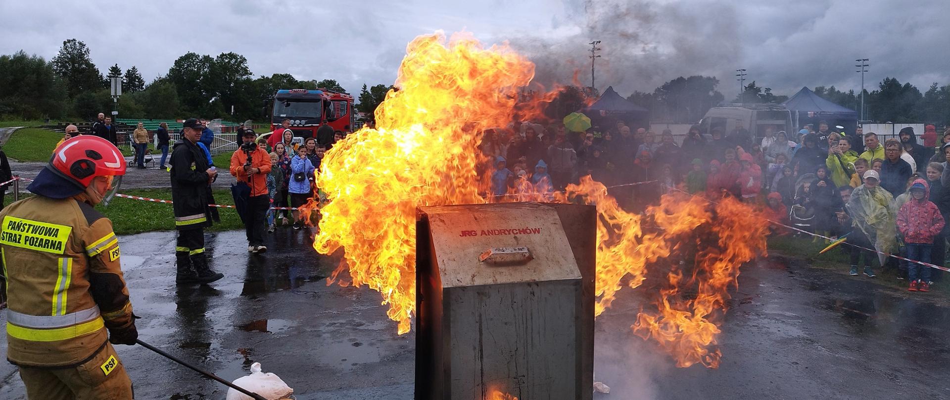 Pokaz wyrzutu oleju. Na pierwszym planie strażak PSP oraz płomienie z palącego się oleju. W tle grupa osób obserwująca pokaz.