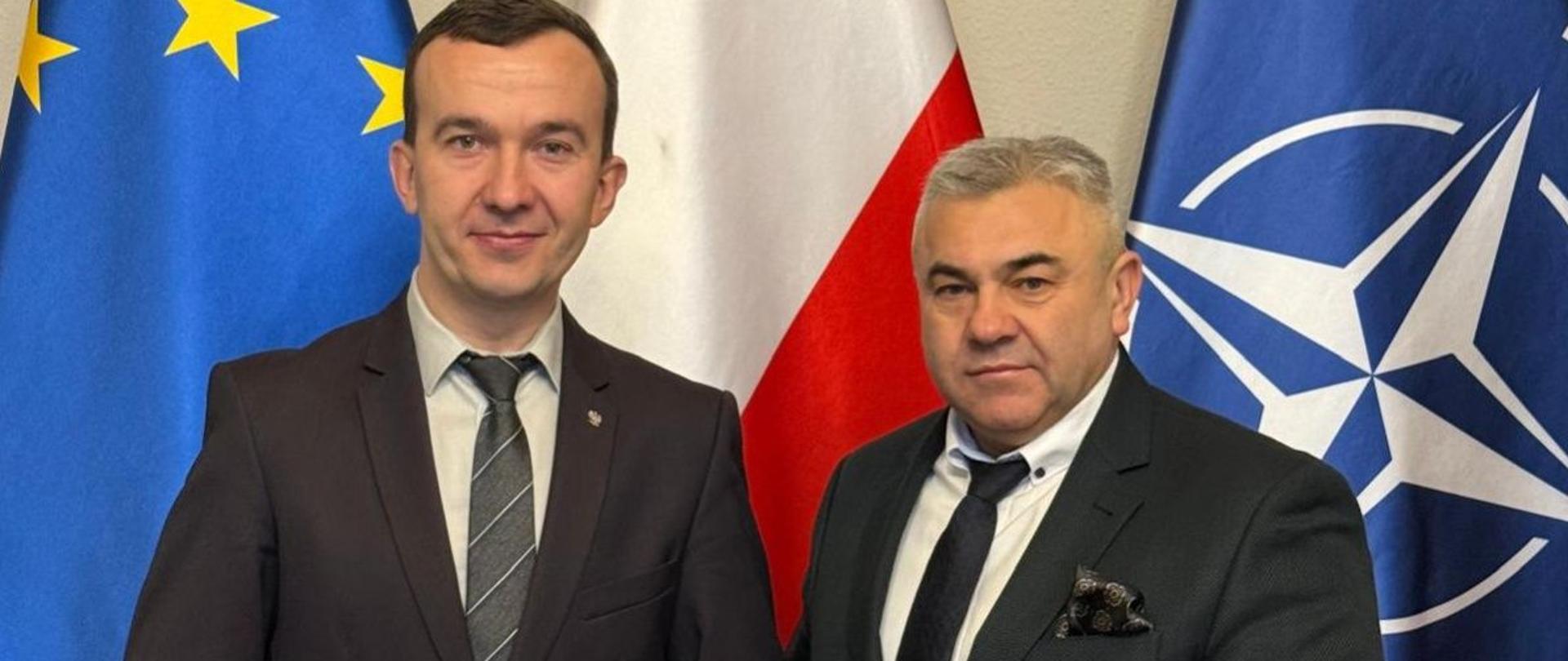 2 mężczyzn w garniturach stoją na tle flag : polskiej, UE i NATO