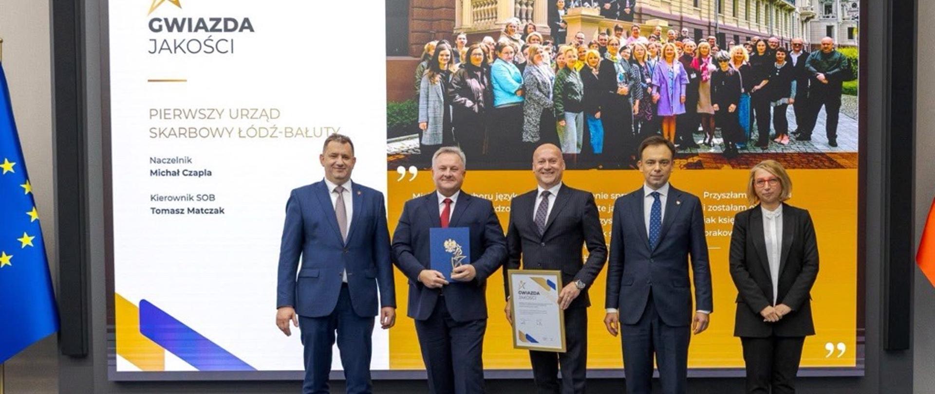 Andrzej Domański, minister finansów i gospodarki, Marcin Łoboda, szef Krajowej Administracji Skarbowej, oraz przedstawiciele Urzędu Skarbowego w Staszowie prezentują dyplom i statuetkę „Gwiazda Jakości”. Na ekranie w tle widoczna jest nazwa urzędu, zdjęcie zespołu oraz fragmenty opinii klientów