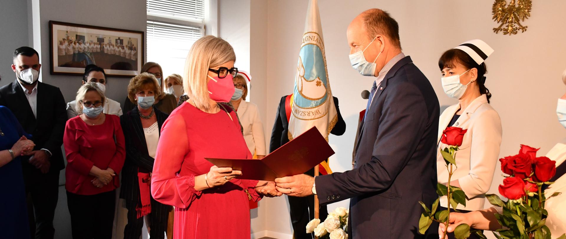 Na zdjęciu znajduje się ubrany w garnitur mężczyzna, który wręcza list gratulacyjny elegancko ubranej kobiecie. 