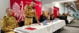 Zdjęcie przedstawia osoby przy stole prezydialnym, przemawia Komendant Wojewódzki, w tle baner reklamowy KP PS{P Starogard Gdański
