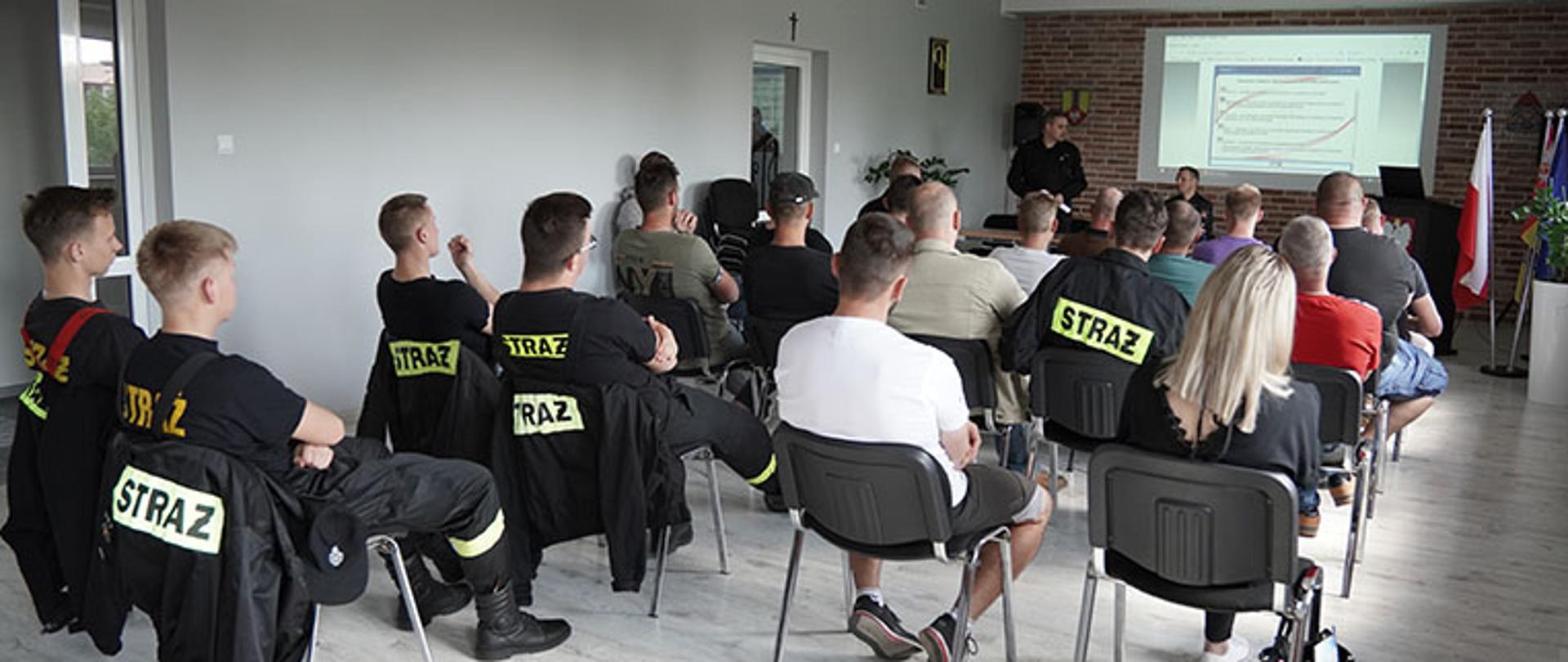 grupa kilkunastu słuchaczy siedzi na krzesłach w pięciu rzędach na sali, przed nimi zastępca komendanta powiatowego ubrany w ubranie sztabowe stoi przed stolikiem, trzyma w dłoni zwiniętą kartkę, drugi strażak ubrany w ubranie sztabowe siedzi za stolikiem, za ich plecami ściana z cegły i ekran projekcyjny na którym wyświetla się strona internetowa z zagadnieniami szkoleniowymi strażaków osp, po ich prawej stronie stoi duża donica z rośliną o drobnych liściach, na ścianie za nimi po bokach ekranu widoczny herb powiatu łowickiego oraz logo Państwowej Straży Pożarnej, w prawym rogu duża donica z rośliną o drobnych liściach oraz stojak a w nim trzy flagi, polski, powiatu łowickiego oraz unii europejskiej, pięć osób na sali ubrana jest w ubranie koszarowe osp