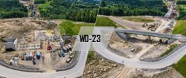 Kolaż dwóch zdjęć lotniczych przedstawiających budowę wiaduktu drogowego oznaczonego napisem „WD-23”. Po lewej stronie widoczny jest obiekt w trakcie budowy z placem robót i sprzętem budowlanym, a po prawej gotowy wiadukt z nową jezdnią. Całość otoczona jest zielonymi wzgórzami i lasami, przez które przebiega budowana trasa.