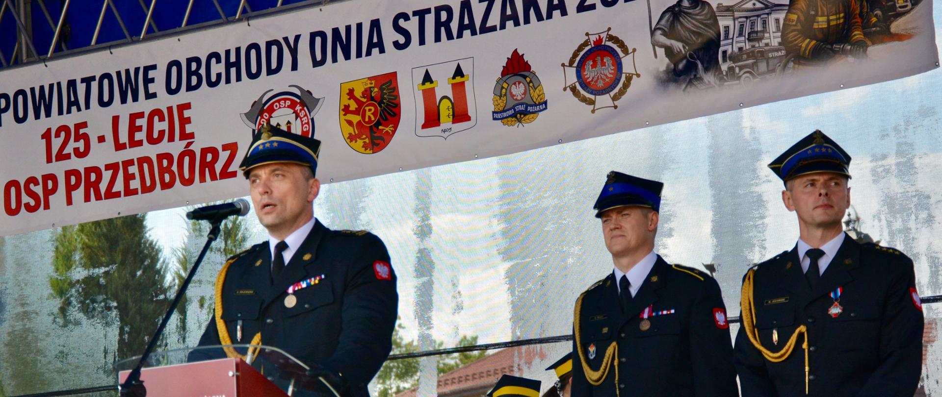 Zbliżenie na mężczyznę w mundurze strażackim, mówiącego do mikrofonu. Obok stoją strażak w mundurze galowym z niebieską czapką. W tle znajduje się transparent z logami jednostek strażackich.