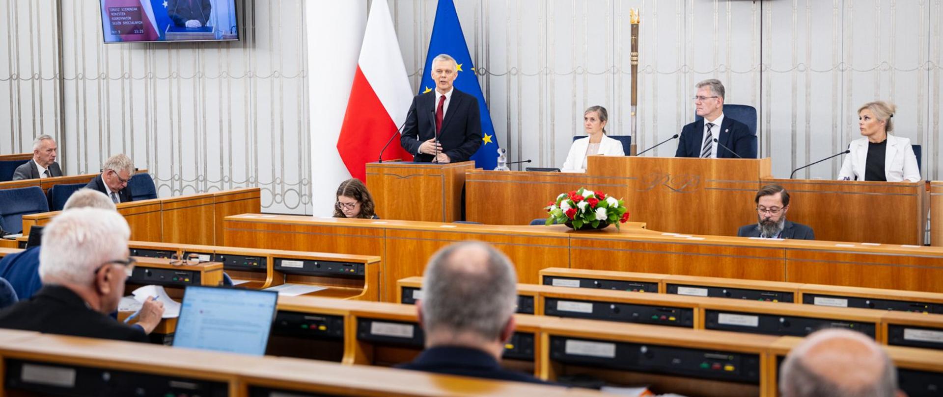 Sala posiedzeń parlamentu. Przy mównicy stoi minister Tomasz Siemoniak przemawiający na tle polskiej i unijnej flagi. Za nim, przy prezydialnym stole, siedzą inni uczestnicy obrad. W pierwszych rzędach widoczni są parlamentarzyści pracujący przy pulpitach z mikrofonami i komputerami. W centralnej części sali znajduje się stół z kwiatami, a wnętrze ma jasne ściany i drewniane elementy wyposażenia.