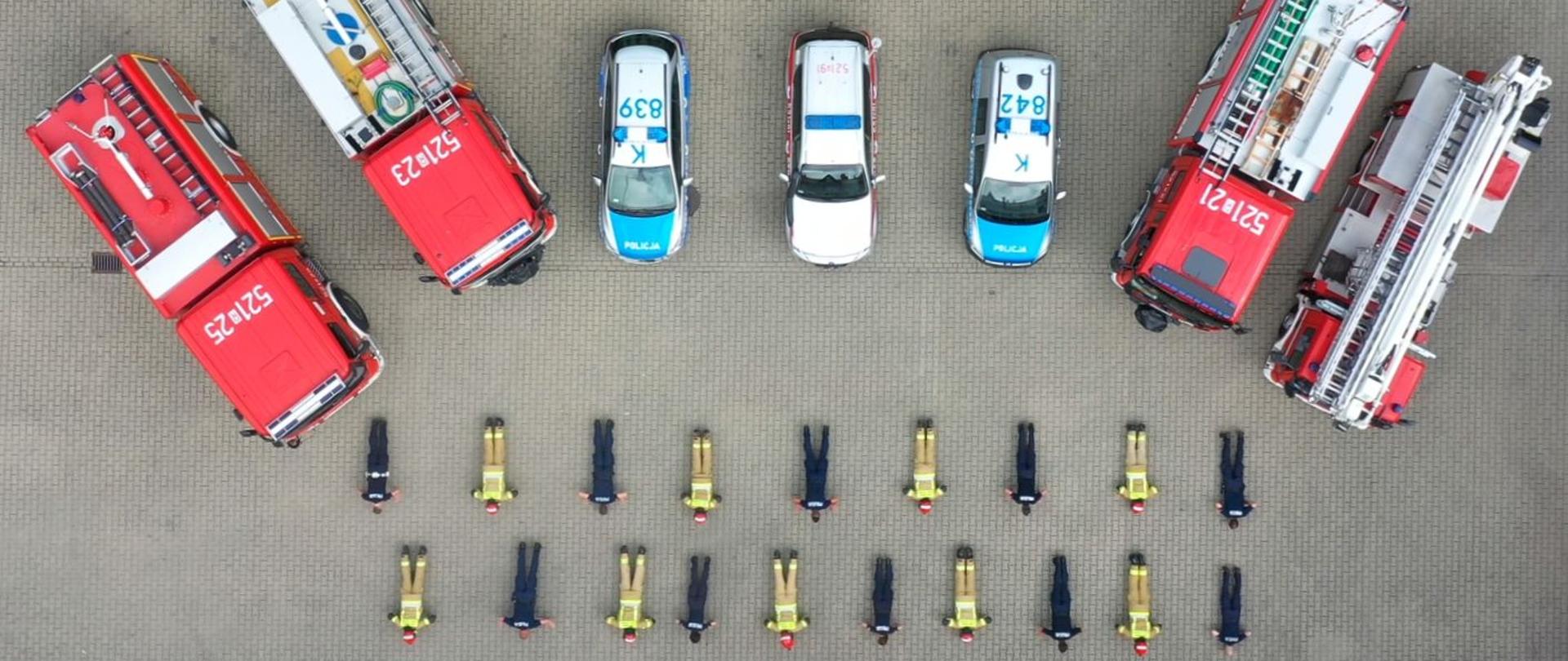 Zdjęcie zrobione z lotu ptaka przedstawia plac wewnętrzny komendy oraz leżących w dwóch szeregach na przemian strażaków i policjantów, którzy robią pompki. U góry zdjęcia ustawione w półkolu osiem samochodów straży pożarnej i policji