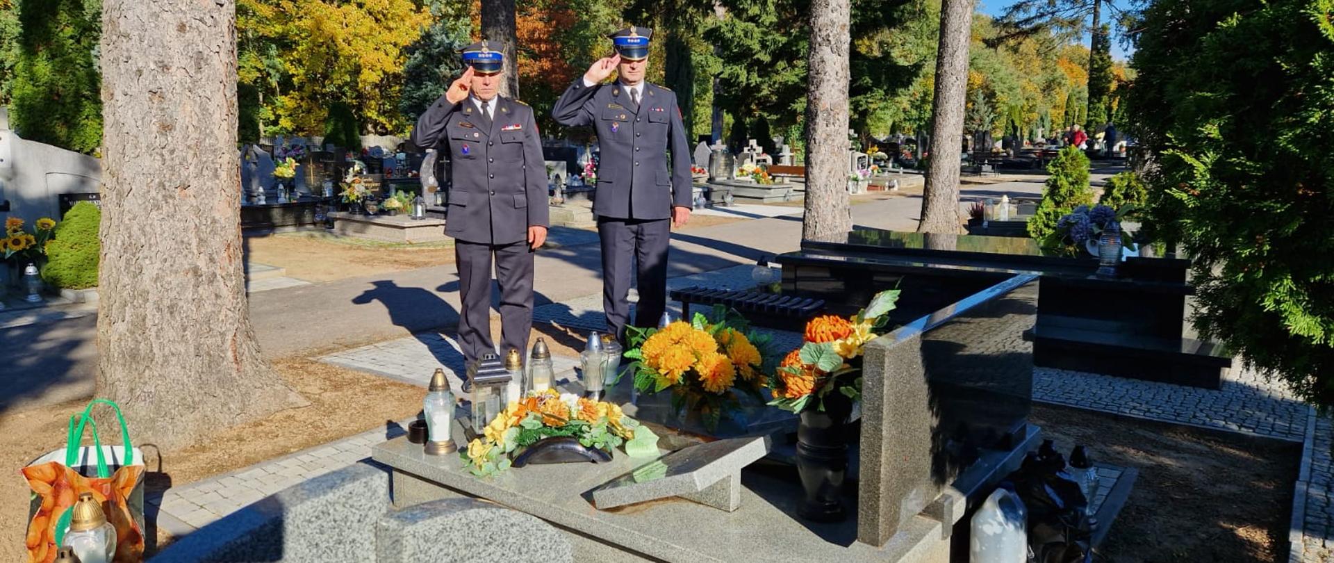 Strażacy oddają honory przed grobem zmarłego funkcjonariusza, na grobowcu kwiaty i znicze 