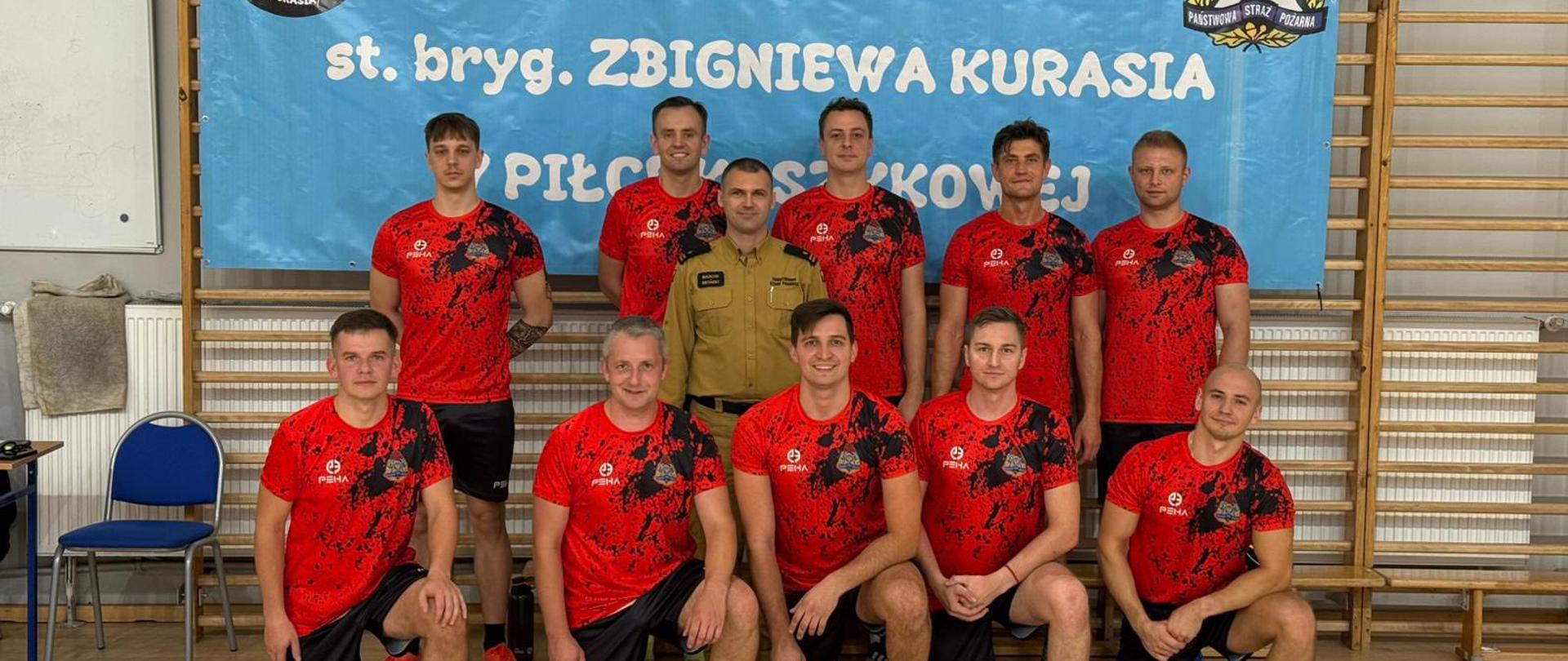Grupa mężczyzn stoi w szeregu na boisku ze sztuczną trawą. Większość z nich ubrana jest w czerwono-czarne stroje sportowe, jeden z mężczyzn jest ubrany w piaskowy strój oficera straży pożarnej. 