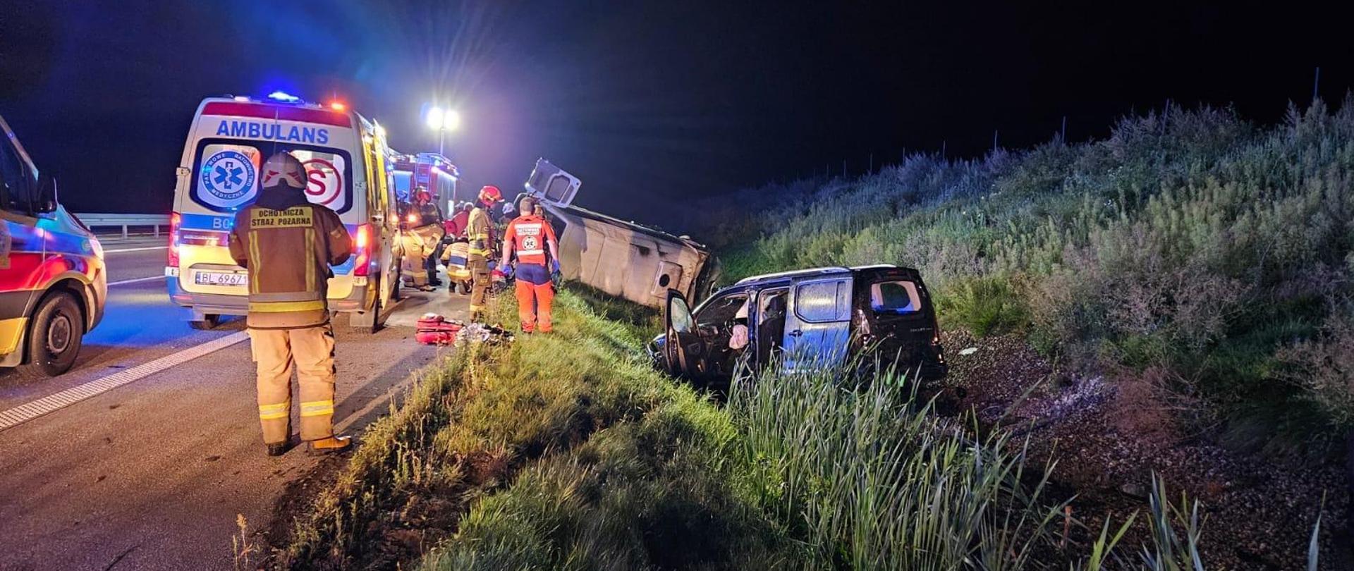 Samochód typu bus oraz samochód osobowy znajdujące się w rowie. Z lewej strony dwie karetki pogotowia i samochód strażacki. W tle strażacy i ratownicy medyczni.