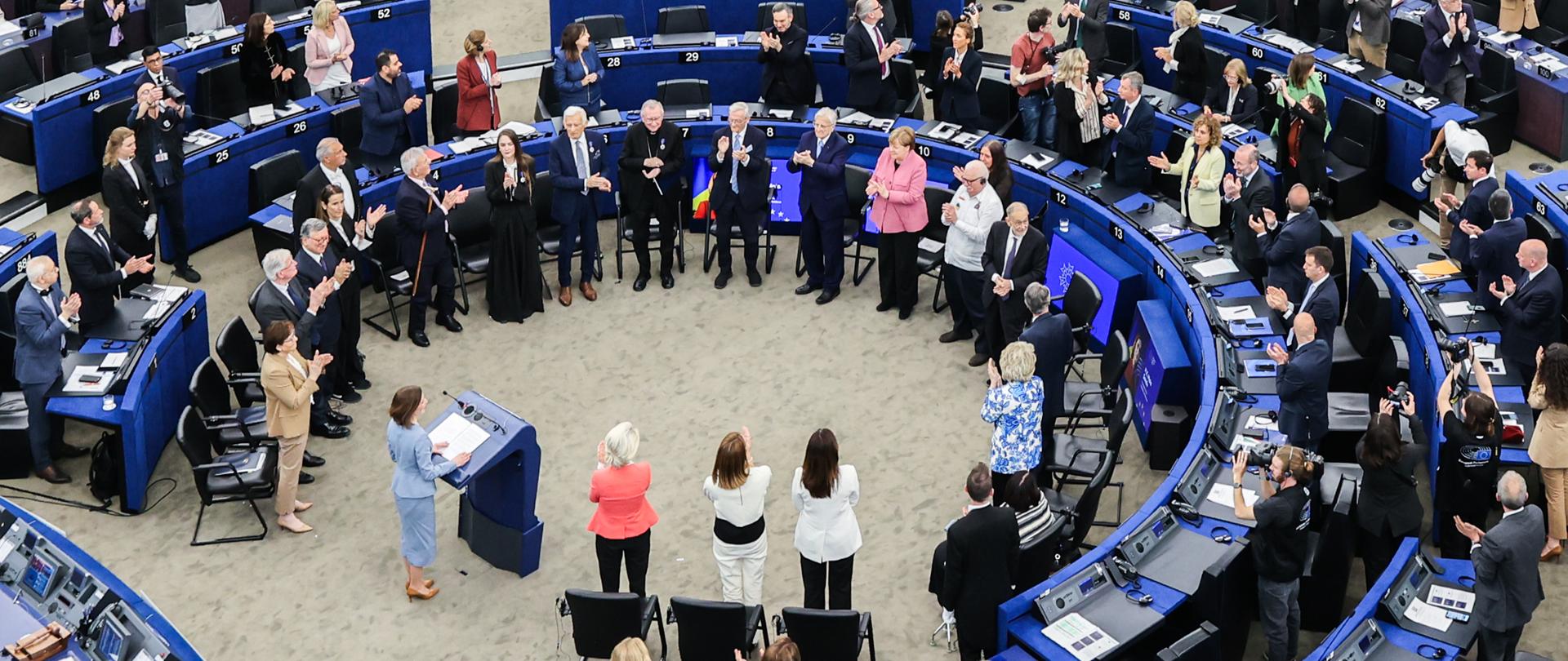 W dużej sali obrad Parlamentu Europejskiego setki uczestników stoją przy półokrągłych rzędach niebieskich stanowisk i biją brawo. W centralnej części sali zgromadziła się grupa osób otoczona przez parlamentarzystów i gości w formalnych strojach.