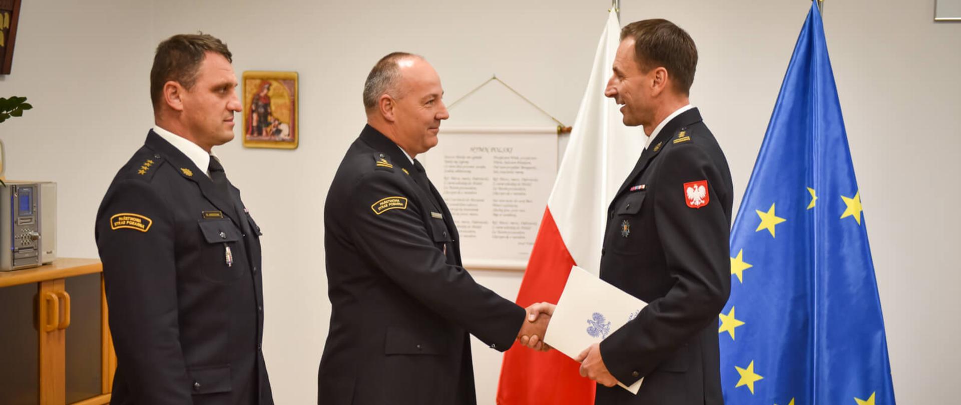 Dwóch mężczyzn w mundurach polskiej służby uściskuje sobie dłonie w geście gratulacji. W tle widać flagę Polski i flagę UE.