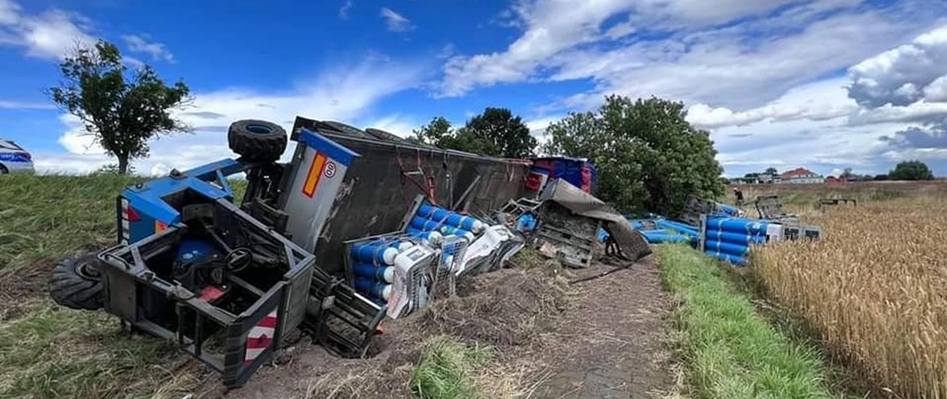 Błękitne niebo z białymi chmurami. Rów a obok pole ze zbożem. Przewrócona czarna naczepa z wózkiem widłowym na przedzie. Obok porozrzucane niebiesko-białe butle gazowe.