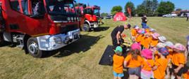 Zdjęcie przedstawia strażaka który pokazuje dzieciom sprzęt będący na wyposażeniu wozów strażackich. Z lewej strony znajdują się wozy strażackie.