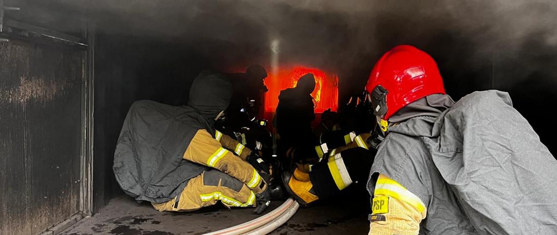 Strażacy PSP uczą strażaków ochotniczej straży pożarnej zachowania w komorze rozgorzeniowej. Strażacy w umundurowaniu bojowych z hełmami i w pełnym zabezpieczeniu realizują ćwiczenia wewnątrz komory rozgorzeniowej. Strażacy siedzą na podłodze a w tle widoczny ogień pożaru. Nad strażakami gęsty szary dym.