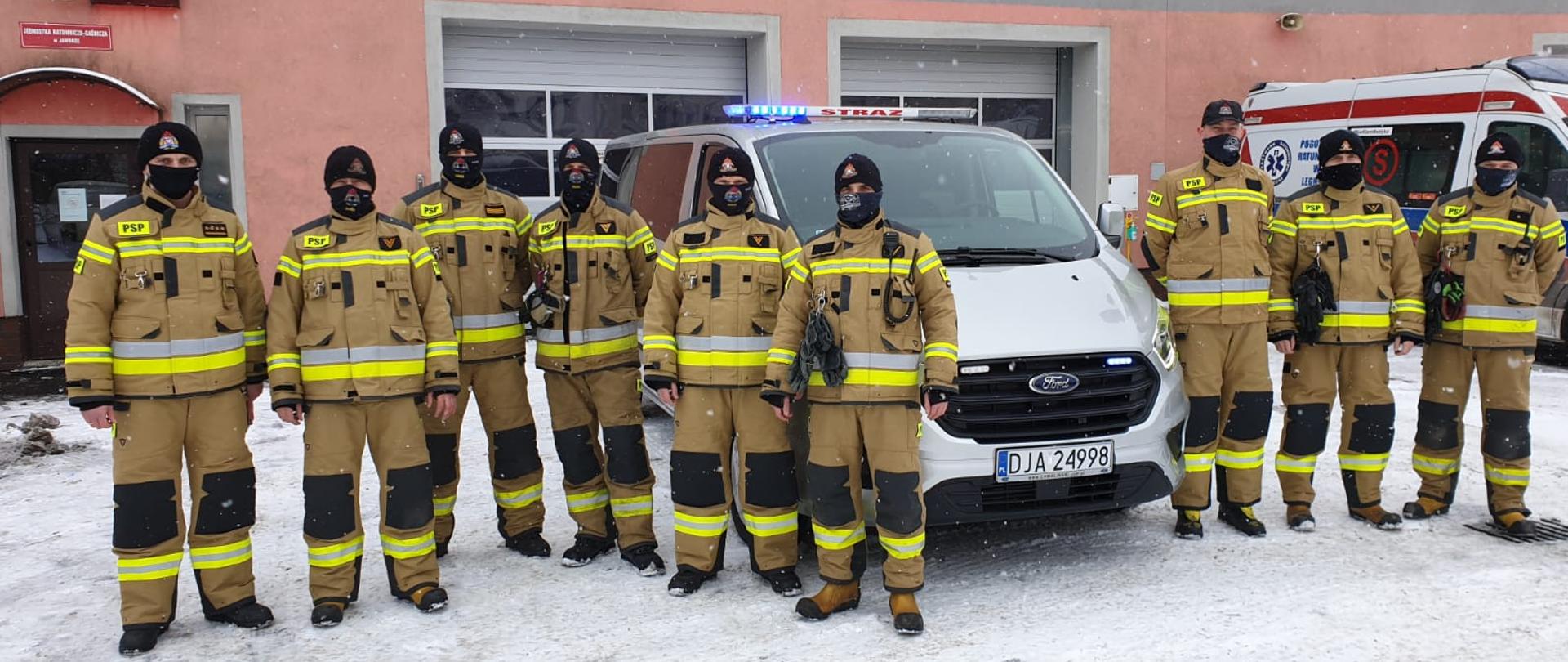 Obraz przedstawia strażaków Komendy Powiatowej Państwowej Straży Pożarnej w Jaworze w nowych ubrania specjalnych. W tle nowy samochód mikrobus oraz budynek komendy.