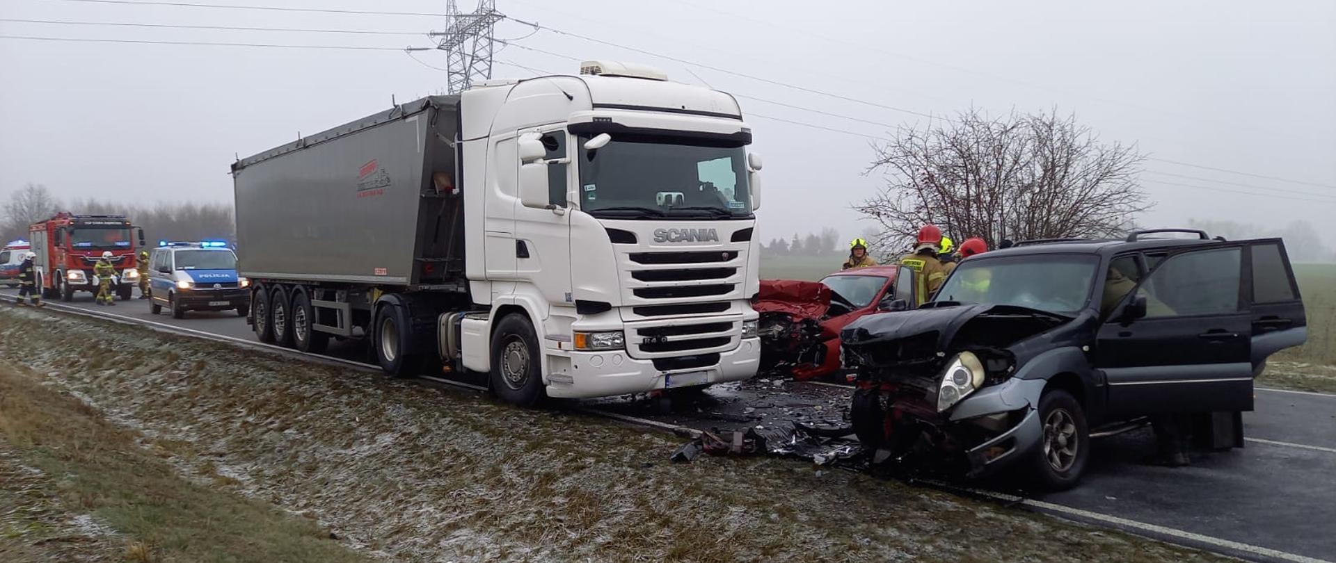 na zdjęciu rozbite samochody pomiędzy nimi strażacy, za nimi radiowóz, samochód pożarniczy