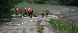 Na zdjęciu widoczni strażacy w czarnych i piaskowych ubraniach koszarowych i w kamizelkach podczas rozwijania węży pożarniczych i organizacji zaopatrzenia wodnego z rzeki. Na wprost widoczna droga z płyt betonowych prowadząca do rzeki.
