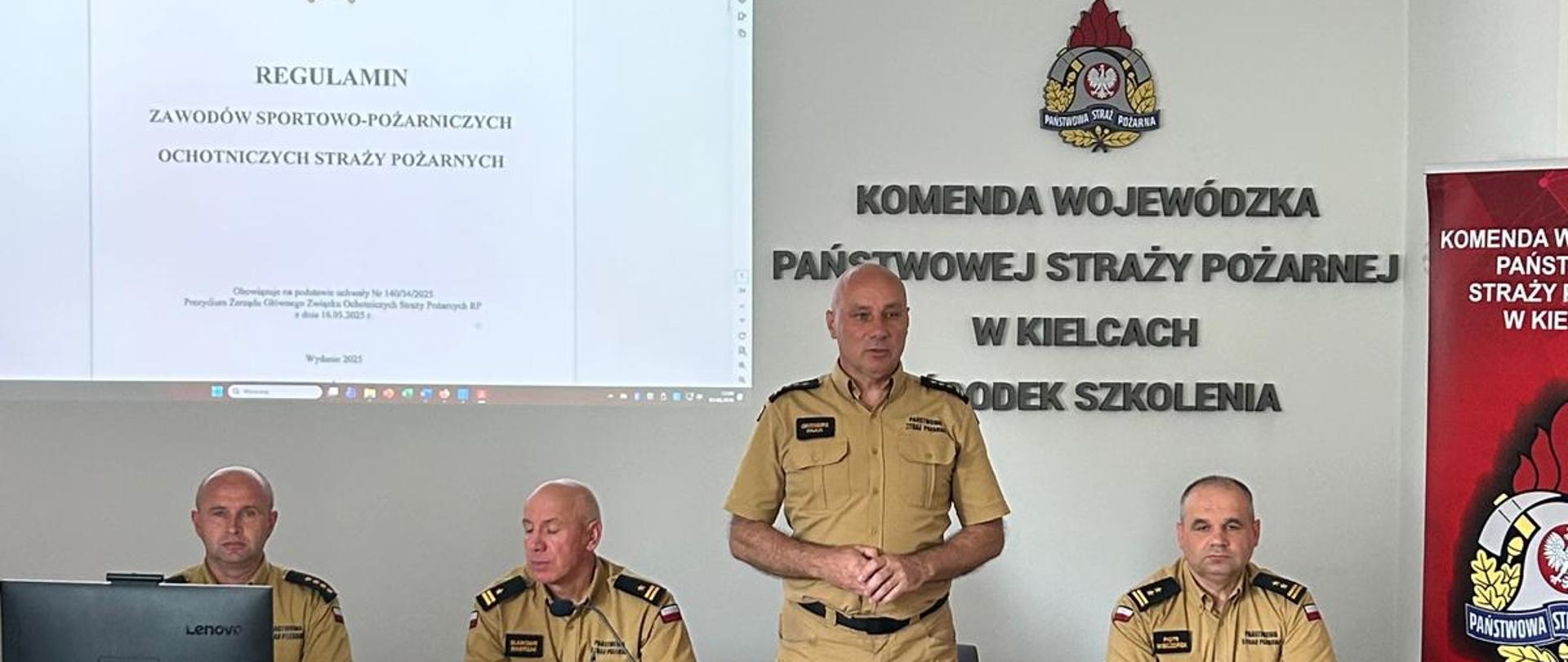 Oficjalne spotkanie w Komendzie Wojewódzkiej Państwowej Straży Pożarnej w Kielcach. Jeden oficer w beżowym mundurze stoi i przemawia, podczas gdy trzech innych siedzi przy stole prezydialnym. W tle widać ekran z prezentacją o regulaminie zawodów strażackich.