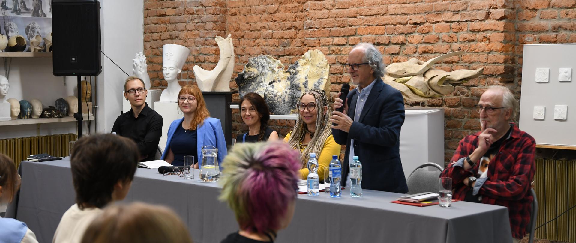 Grupa sześciu osób siedzi przy stole przykrytym szarym obrusem podczas spotkania z publicznością. Jeden z mężczyzn, stojący przy stole, mówi do mikrofonu. Na ścianie za uczestnikami widać ceglaną fakturę oraz różne rzeźby i popiersia. Przed stołem siedzi publiczność, częściowo widoczna od tyłu.