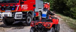 Na zdjęciu widoczny na piaskowej drodze leśnej quad którym porusza się strażak w ubraniu czarnym dowódczo-sztabowym i biało niebieskiej kamizelce po jego prawej stronie las iglasty a po lewej czerwony samochód pożarniczy.