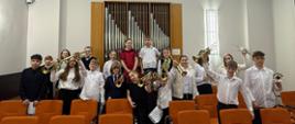 Na zdjęciu widoczna jest grupa uczniów klasy trąbki pozujących w sali koncertowej. Dzieci i młodzież stoją między rzędami pomarańczowych krzeseł, trzymając w rękach trąbki, część z nich unosi instrumenty w górze. Uczniowie ubrani są w stroje koncertowe w jasnych i ciemnych kolorach. W tle znajduje się prospekt organowy z piszczałkami oraz jasne ściany sali. Zdjęcie przedstawia radosny moment po wspólnym występie muzycznym.