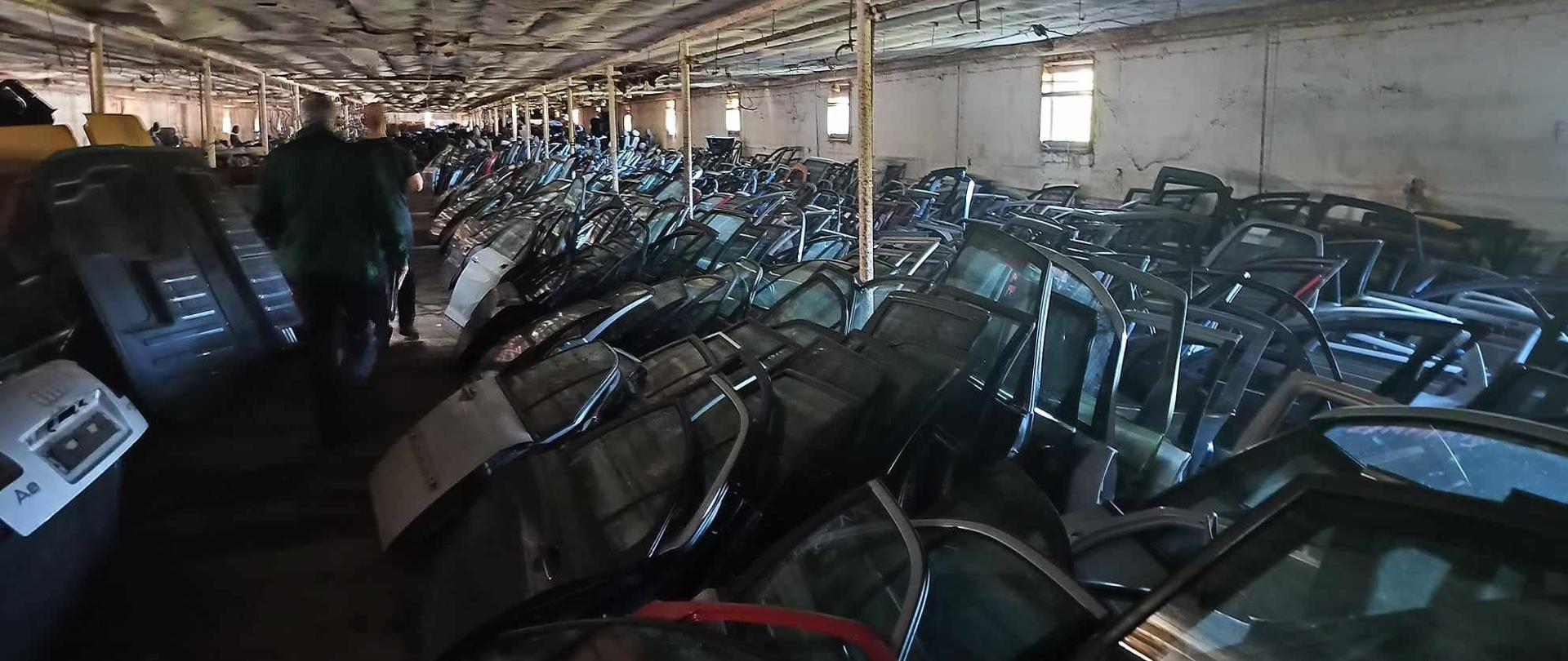 Widoczny magazyn z zamieszczonymi częściami do samochodu oraz ukazanymi dwoma mężczyznami obróconymi tyłem.