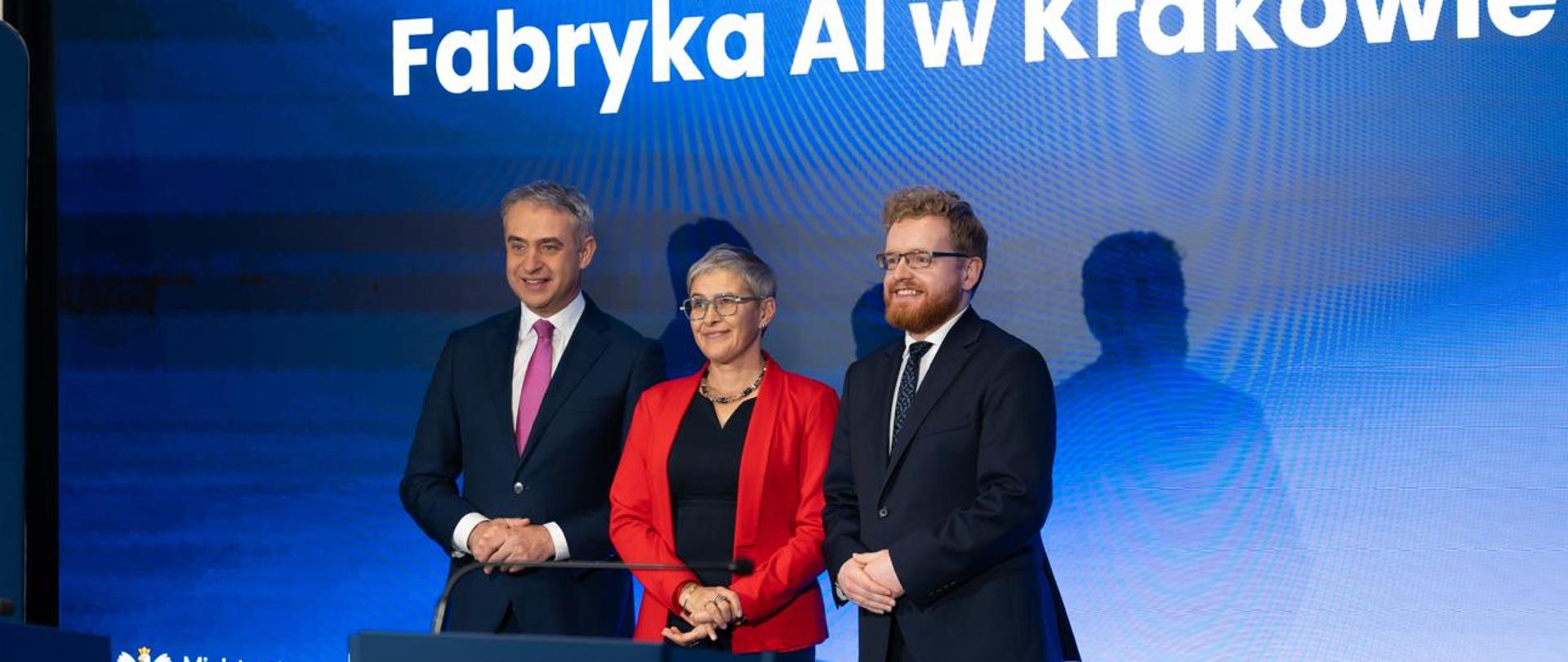 Za mównicą stoi wiceminister Mrówczyńska w czerwonej marynarce między dwoma mężczyznami w czarnych garniturach, za nimi na ścianie wielki ekran z napisem "Fabryka AI w Krakowie" na niebieskim tle.