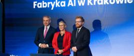 Za mównicą stoi wiceminister Mrówczyńska w czerwonej marynarce między dwoma mężczyznami w czarnych garniturach, za nimi na ścianie wielki ekran z napisem "Fabryka AI w Krakowie" na niebieskim tle.