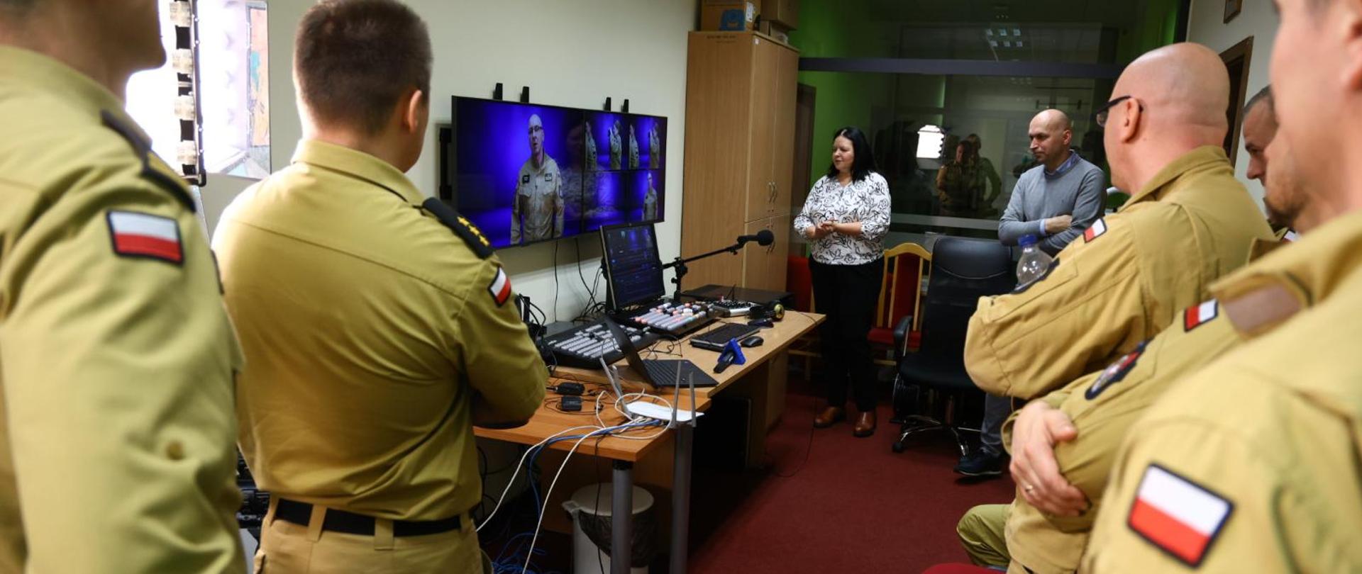 W studiu telewizyjnym kilkoro strażaków stoi i spogląda na monitory wiszące na ścianie. Tam trwa projekcja wcześniejszych nagrań z ich udziałem. Widać konsoletę mikrofony, w tle szklana ściana.
