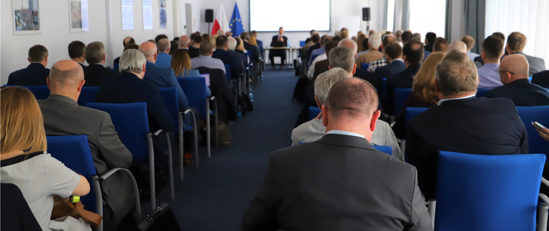 Sala konferencyjna. Kilkadziesiąt osób siedzi i słucha prezentacji. Na końcu sali siedzi prowadzący spotkanie mężczyzna z mikrofonem. Za nim ekran z prezentacją - treść niewidoczna. Obok flagi polska i unijna.